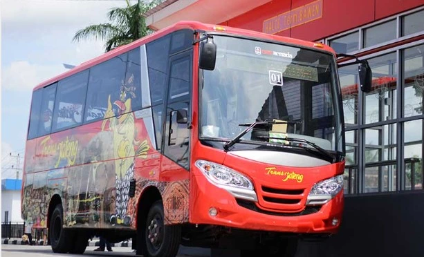 Foto eksterior bus medium Trans Jateng berwarna merah terang dengan branding logo dan maskot wayang, terparkir di depan Halte Terminal Bawen yang modern.