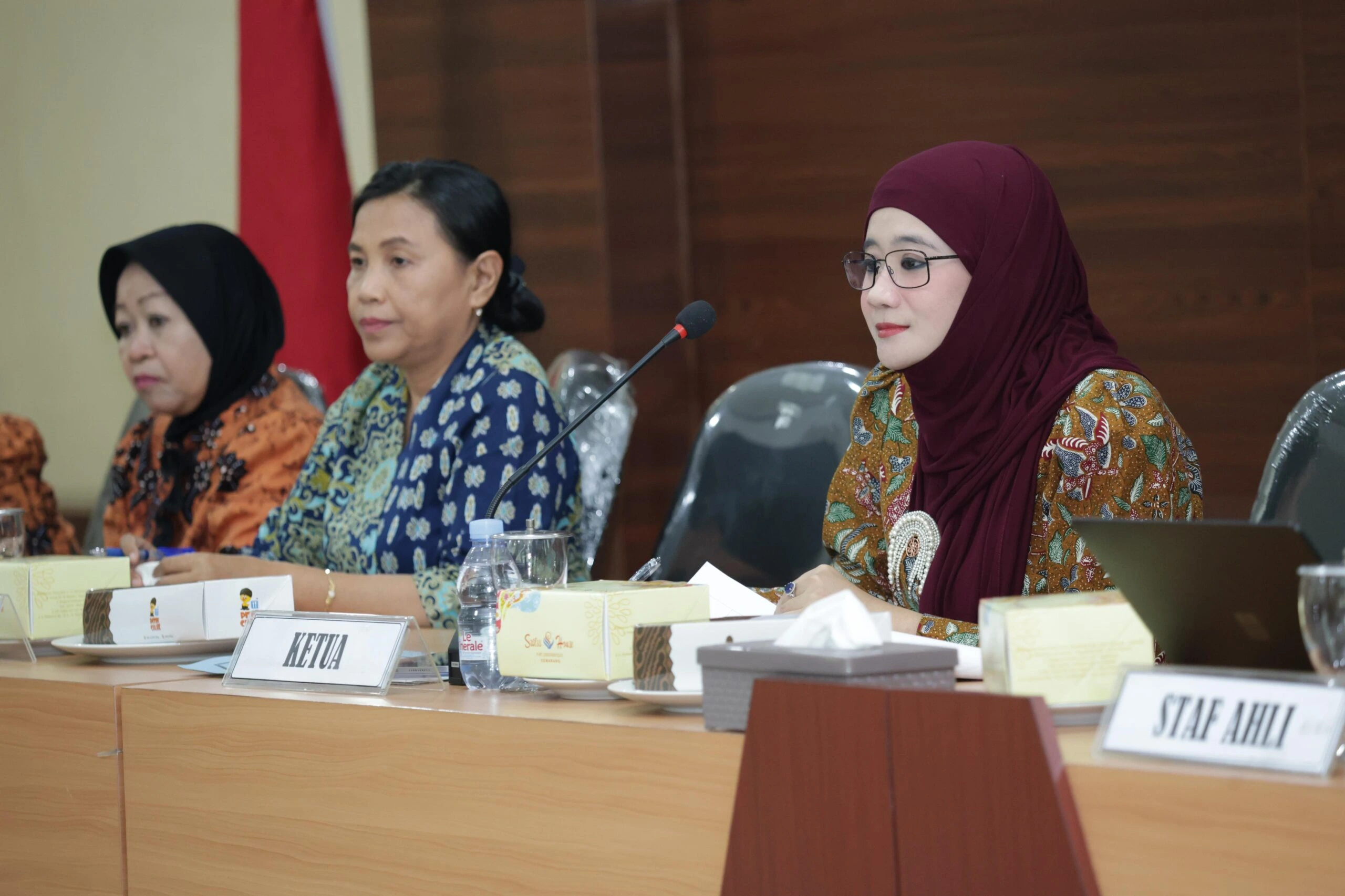 Tiga pejabat wanita di meja rapat pleno, wanita di tengah berlabel 'KETUA' dan wanita di kanan berlabel 'STAF AHLI', dengan bendera Merah Putih di latar belakang.