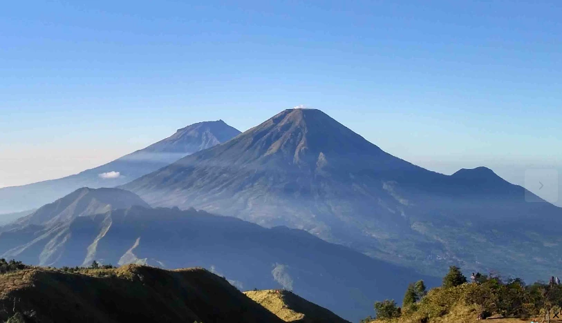 emandangan lanskap pegunungan Jawa Tengah yang luas dengan dua puncak gunung berapi kembar agung yang menonjol di bawah langit biru cerah. Di latar depan, sebuah bukitkamp yang lebih dekat memiliki beberapa tenda kecil berwarna-warni dan sekelompok kecil pendaki yang sedang beraktivitas. Lapisan kabut tipis mengisi lembah di antara pegunungan.
