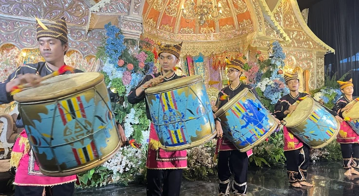 Lima pria berpakaian adat menabuh alat musik tambur berukuran besar di depan pelaminan bernuansa emas pada resepsi pernikahan adat Minang di Pesawaran, Lampung.