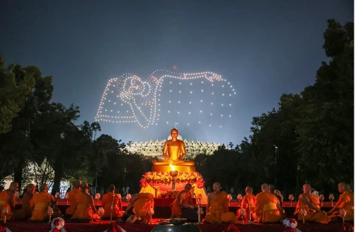 Barisan biksu berjubah jingga bermeditasi khusyuk di depan patung Buddha emas, berlatar belakang siluet candi dan indahnya pendaran cahaya Lampion Waisak Borobudur di langit malam Magelang.