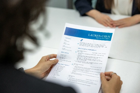 Tampilan dari belakang seorang wanita memegang dan meninjau dokumen CV profesional modern dengan aksen biru bertuliskan nama LAUREN CHEN di atas meja selama sesi wawancara kerja.