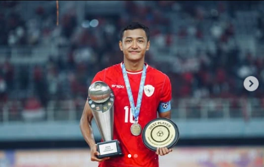 Dony Tri Pamungkas, pemain asli Boyolali Jawa Tengah, tersenyum bangga memegang trofi piala dan plakat Best Player mengenakan jersey merah Timnas Indonesia dengan latar belakang stadion.