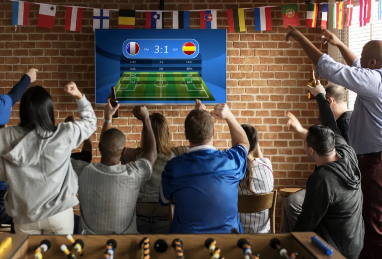 Sekelompok suporter sepak bola bersorak gembira merayakan gol sambil menonton tayangan pertandingan di layar televisi dalam sebuah kafe bernuansa dinding bata.