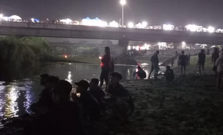 Warga bersantai di tepi sungai remang saat tradisi Kungkum di Tugu Suharto, sementara jembatan di atasnya terang benderang dipenuhi deretan tenda wisata kuliner Semarang malam pada perayaan Malam 1 Suro.