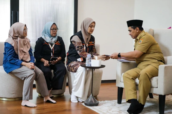 "Wakil Gubernur Jawa Tengah Taj Yasin Maimoen (kanan) duduk di kursi berlengan, mengenakan seragam dinas cokelat dan peci, menerima sebuah perangkat tablet/HP dari seorang petugas sensus wanita (tengah) yang mengenakan rompi BPS dan hijab. Dua wanita lain juga duduk di sebelah kiri dalam interaksi ini."