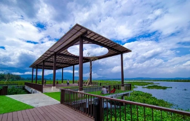 Pemandangan fasilitas anjungan kayu modern di tepi danau Wisata Rawa Pening, Kabupaten Semarang, dengan wisatawan menikmati pemandangan air, tanaman eceng gondok, dan latar belakang pegunungan di bawah langit biru.