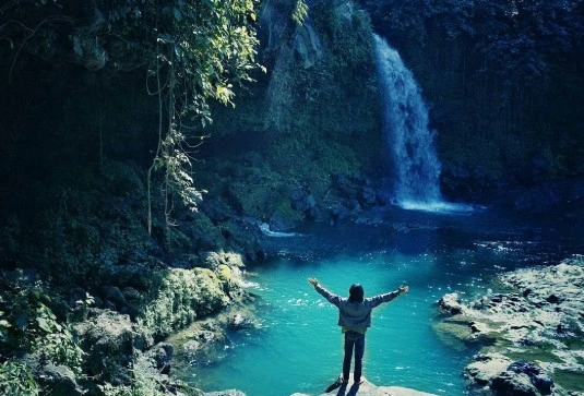 Seorang wisatawan berdiri dengan tangan terangkat di depan Curug Sumba di Purbalingga, Jawa Tengah, dengan latar belakang air terjun dan kolam biru jernih.