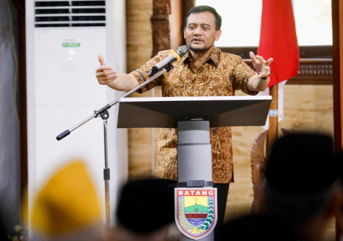 Ahmad Luthfi, Gubernur Jawa Tengah, berjanggut tipis, mengenakan kemeja batik cokelat, berdiri di belakang podium berlogo Batang. Tangannya terangkat tinggi dan terbuka dalam gerakan ekspresif saat berbicara ke mikrofon. Di latar belakang terdapat bendera Merah Putih dan unit AC vertikal. Siluet buram penonton di latar depan.