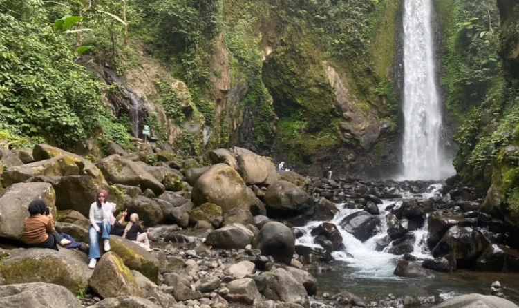 Sekelompok pengunjung bersantai di atas bebatuan menikmati pesona wisata Blado berlatar belakang kemegahan air terjun Curug Genting terbaru, sebuah destinasi wisata alam Batang yang kini aman karena dilengkapi sistem mitigasi bencana wisata.