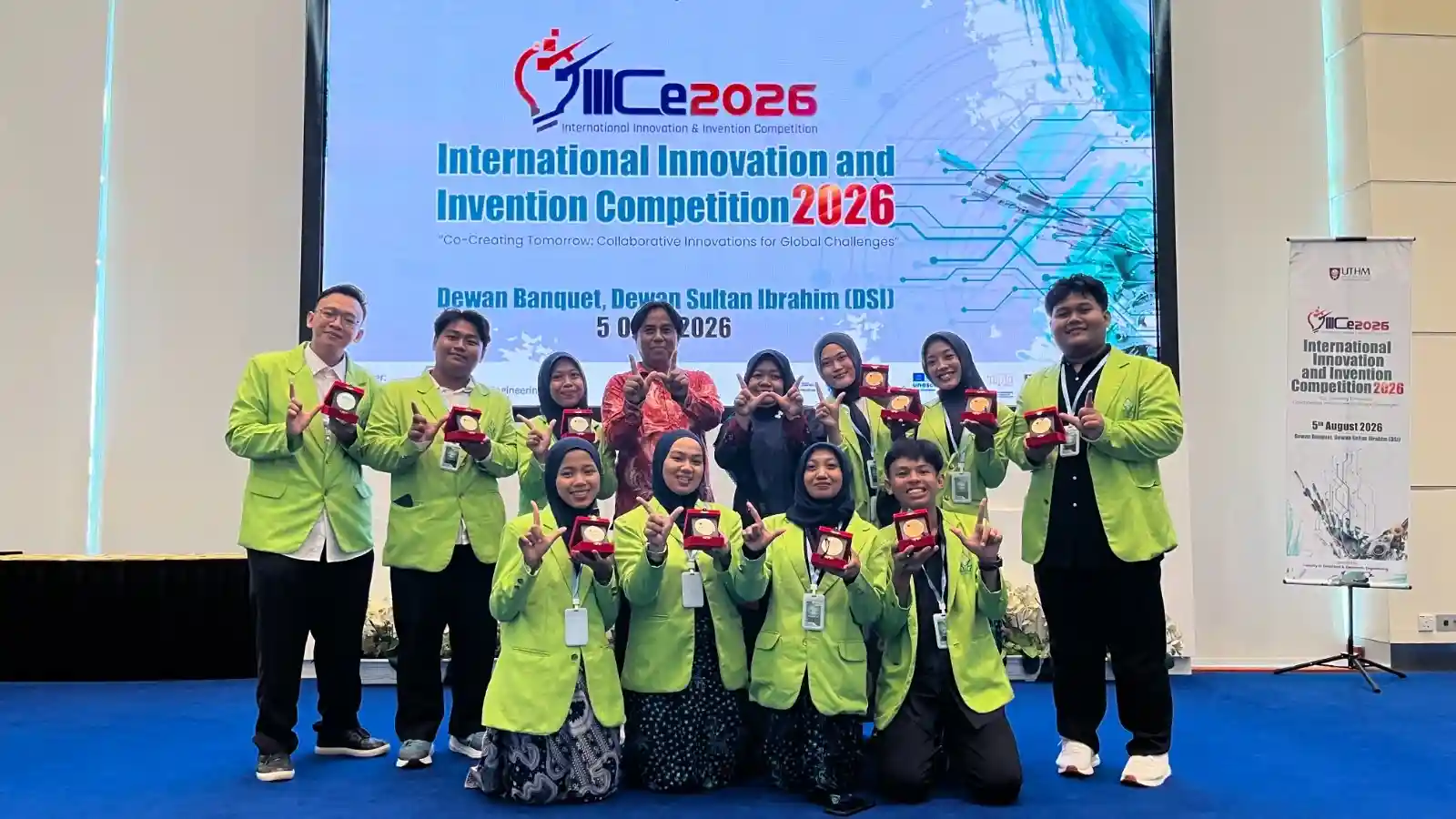 Delegasi mahasiswa FST UIN Walisongo Semarang mengenakan jas almamater hijau memamerkan medali penghargaan di atas panggung ajang inovasi internasional IIICe 2026 UTHM Malaysia.