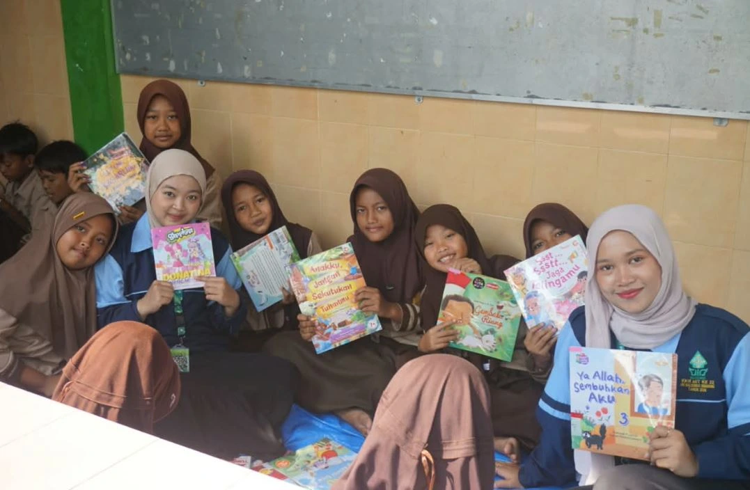 Mahasiswi KKN UIN Walisongo berfoto bersama siswi perempuan SDN Babat 2 Demak yang tersenyum memegang buku cerita bergambar sambil duduk lesehan di atas terpal biru.