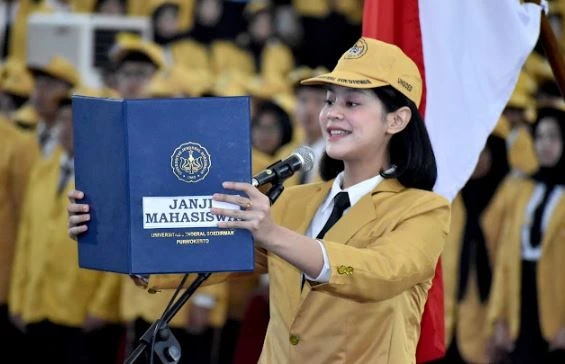 Seorang mahasiswi perwakilan mahasiswa baru Unsoed 2026 mengenakan jas almamater kuning sedang membacakan Janji Mahasiswa di depan mikrofon saat Sidang Terbuka Senat PMB di Graha Widyatama Purwokerto.