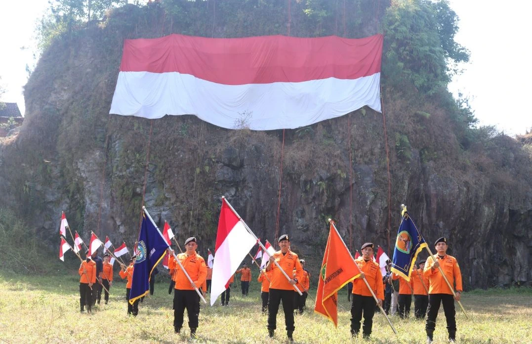Personel SAR Semarang berseragam oranye berdiri tegap membawa panji-panji kebanggaan dengan latar belakang bendera merah putih raksasa yang terbentang di dinding cadas Tebing Djaro Kendal.