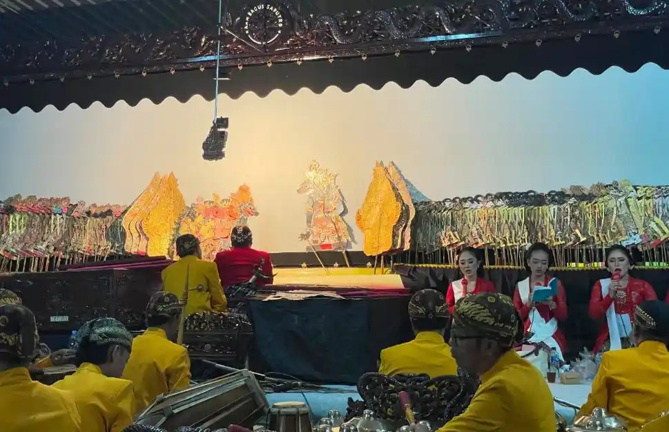 Siluet gunungan wayang kulit di kelir dengan pesinden berkebaya merah dan penabuh gamelan berbaju kuning dalam perayaan tradisi Merti Dusun di Kadipiro Tuntang.