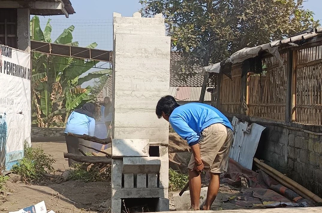 eorang pemuda berbaju biru membungkuk untuk memeriksa bagian bawah tungku rocket stove dari semen di sebuah halaman desa, sementara beberapa warga terlihat di latar belakang.