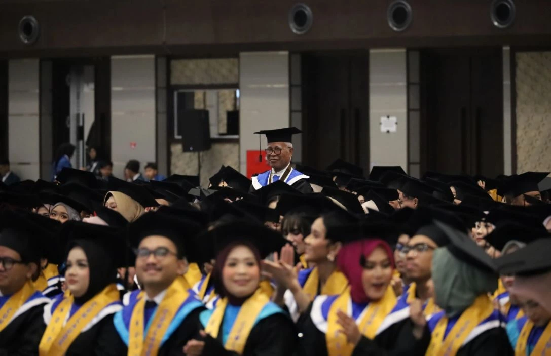 Dr JLM Siahaan, wisudawan Undip tertua berusia 73 tahun, mengenakan toga dan berdiri tersenyum karismatik di tengah kerumunan wisudawan muda yang sedang duduk saat prosesi kelulusan di Muladi Dome.