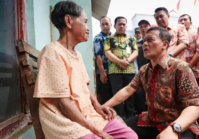 Ahmad Luthfi dengan kemeja batik merah berlutut dan memegang erat tangan Ibu Darmi, seorang lansia yang duduk di kursi kayu di Pekalongan, memastikan pemulihan hak bantuan sosial pasca kasus penyalahgunaan KTP.