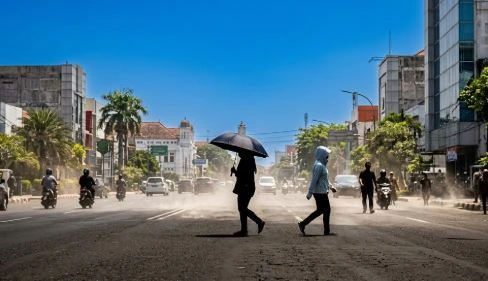 Warga berjalan melintasi aspal jalanan Kota Semarang yang berasap akibat cuaca panas ekstrem, berlindung menggunakan payung hitam dan jaket tudung di bawah sengatan matahari terik.