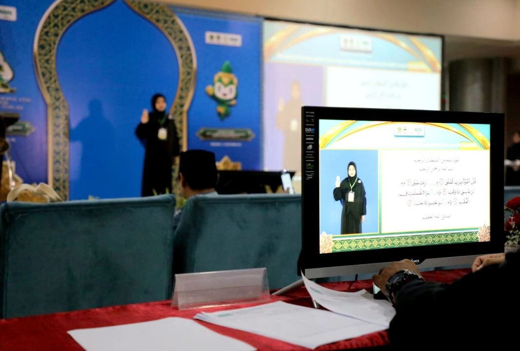 Sudut pandang dari meja dewan hakim menunjukkan sebuah layar monitor yang menampilkan peserta perempuan berhijab hitam mempraktikkan cara tunarungu membaca Al-Qur'an dengan isyarat tangan, bersanding dengan teks ayat suci. Di latar belakang yang sedikit buram, peserta tersebut tampak sedang tampil langsung di atas panggung MTQ Nasional XXXI 2026 berornamen biru dan emas.