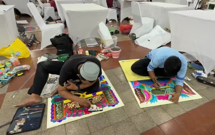 Dua seniman duduk bersila di lantai sedang fokus mengerjakan kanvas Seni Lukis Islami pada kompetisi Lomba Kaligrafi MTQ 2026 di Auditorium Undip Semarang.