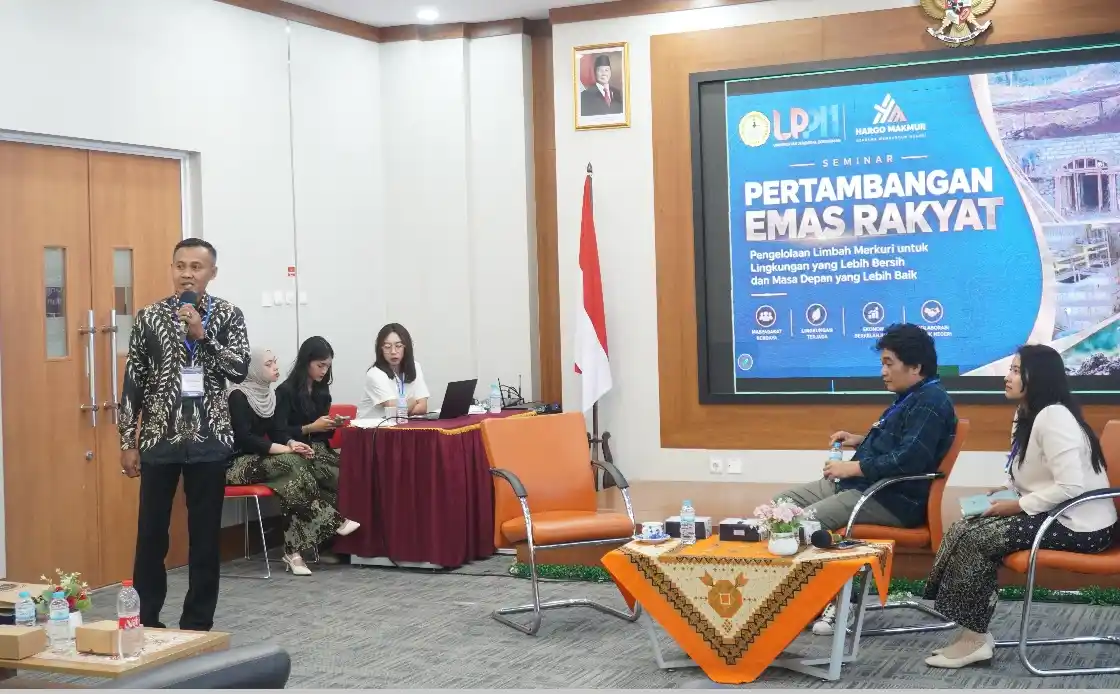 Seorang narasumber pria berbatik berdiri memegang mikrofon memberikan pemaparan di depan layar bertuliskan Seminar Pertambangan Emas Rakyat di ruangan LPPM Unsoed.