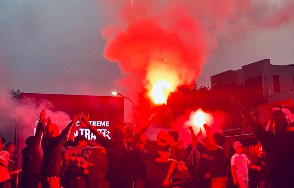 Kerumunan suporter sepak bola PSIS Semarang (Snex) dengan tangan terangkat menyalakan suar merah besar di luar ruangan saat senja di depan tanda bertuliskan 'SEMARANG EXTREME ULTRA'
