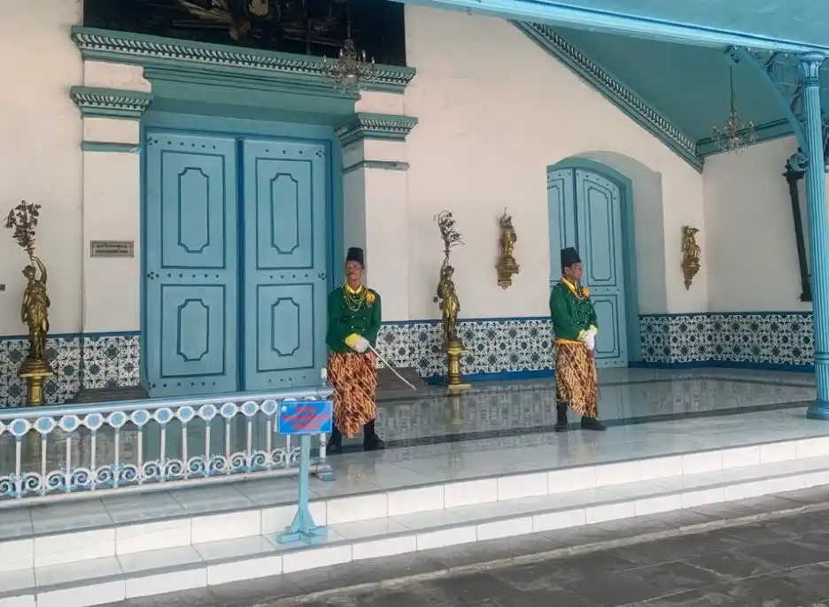 Dua abdi dalem Keraton Kasunanan Surakarta berseragam hijau dan kain batik berdiri menjaga area depan pintu biru megah, melengkapi pesona sejarah dalam rute wisata Solo.