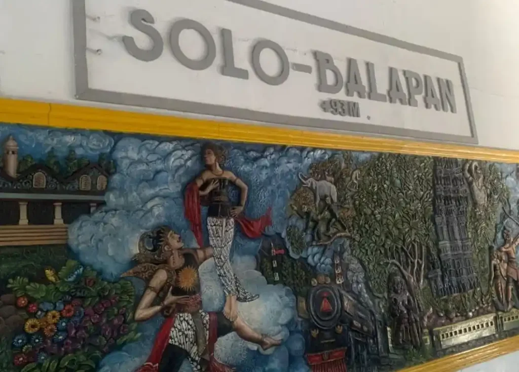 Papan penanda Stasiun Solo Balapan bernuansa klasik dengan ukiran relief tokoh pewayangan dan elemen lokomotif yang menyambut kedatangan pelancong saat memulai wisata Solo.