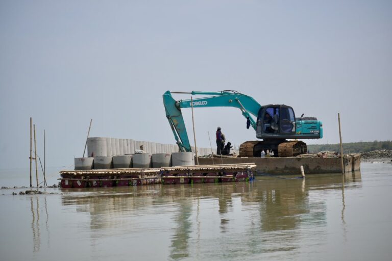 Satu unit alat berat ekskavator berwarna hijau toska berdiri di atas tongkang terapung di laut sedang menyusun jajaran bis-bis beton struktur Hybrid Sea Wall Sayung di pesisir Demak.